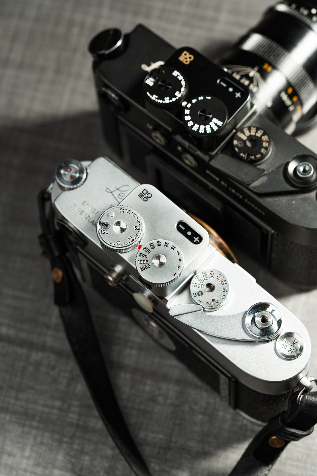 Two cameras on a table shot from above. A chrome and a black Doomo Meter D hot shoe light meters attached to top of both.