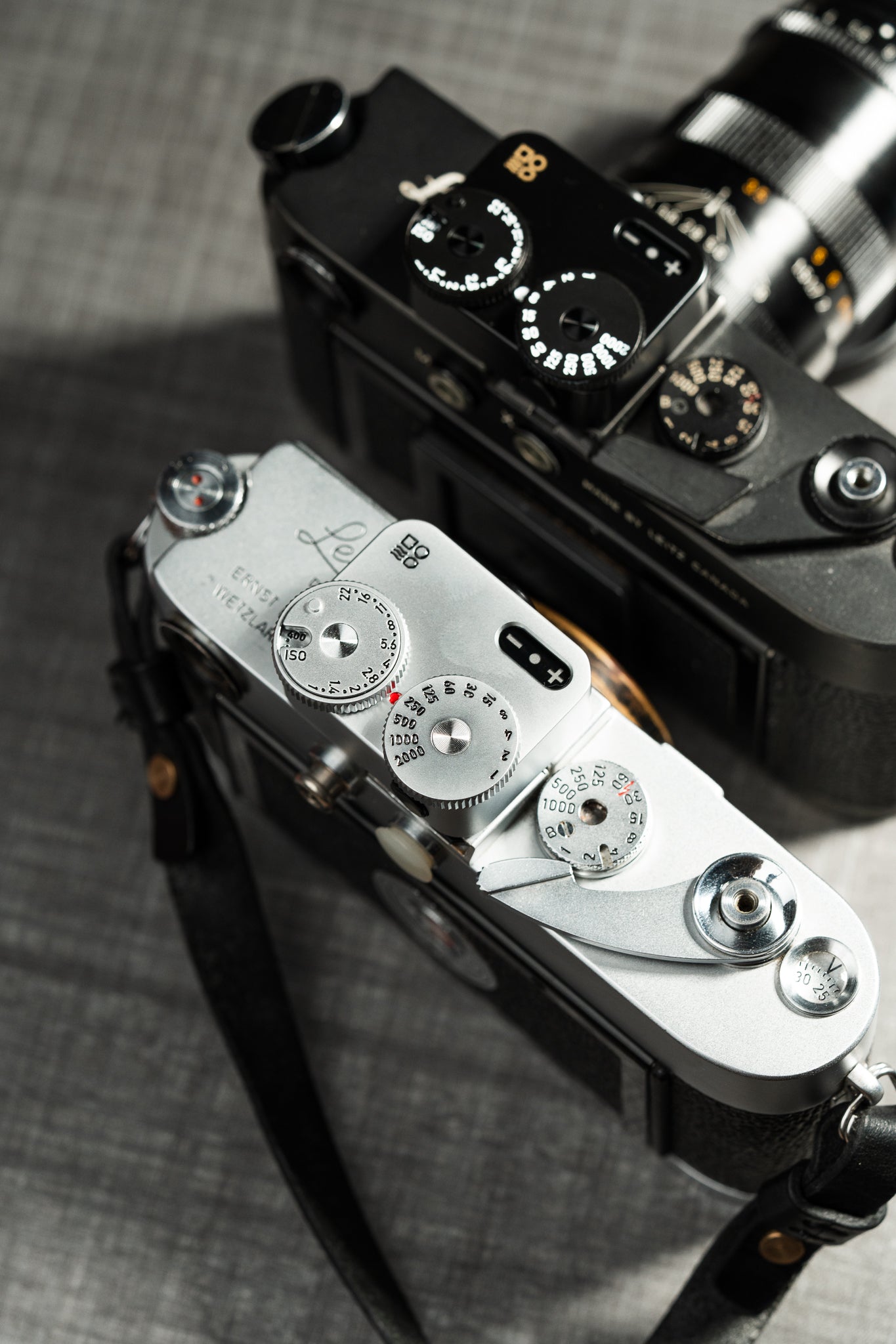 Two cameras on a table shot from above. A chrome and a black Doomo Meter D hot shoe light meters attached to top of both.