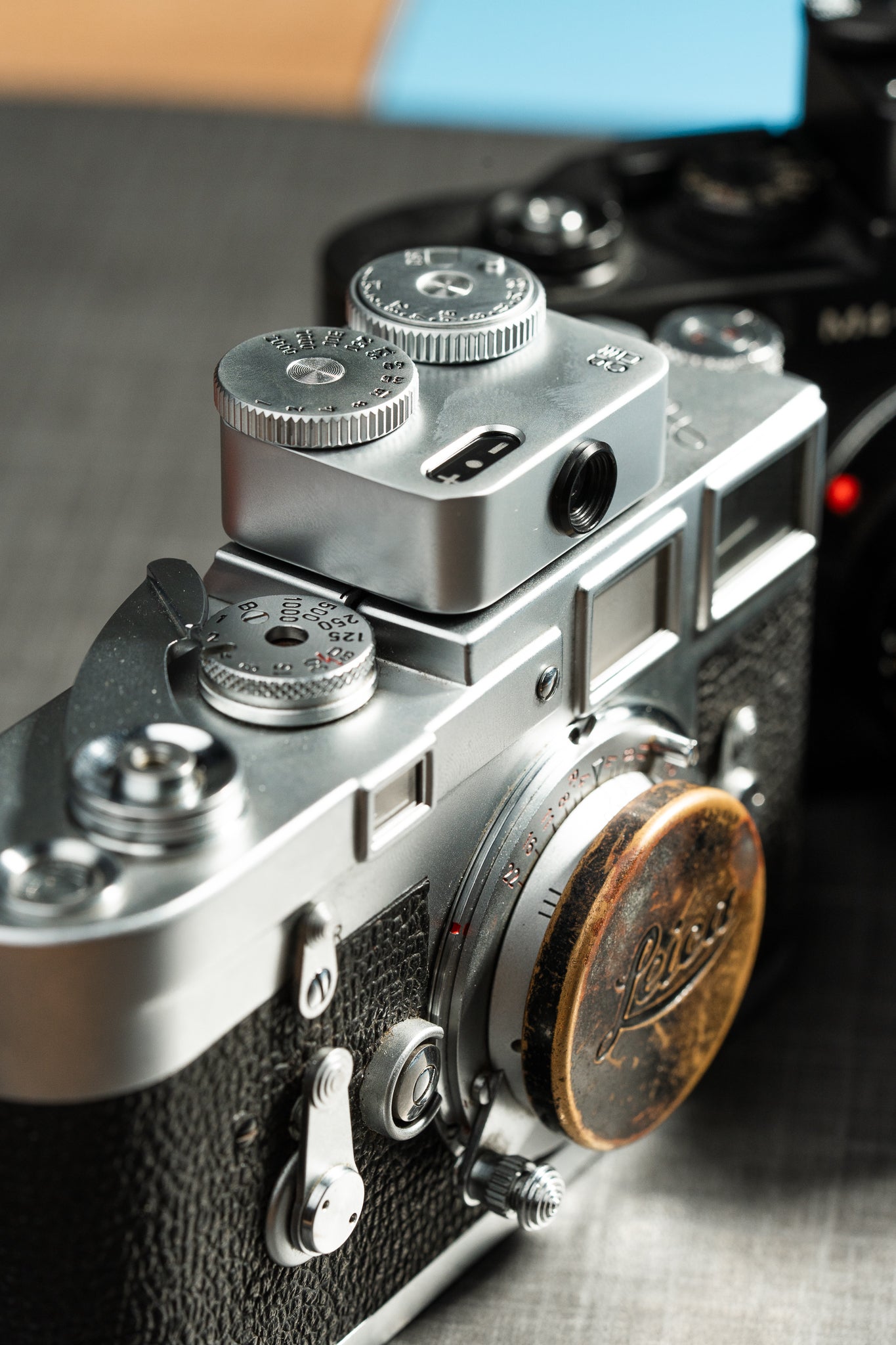 A close-up shot from the side of a camera on a table. A chrome Doomo Meter D hot shoe light meter attached on top of it.