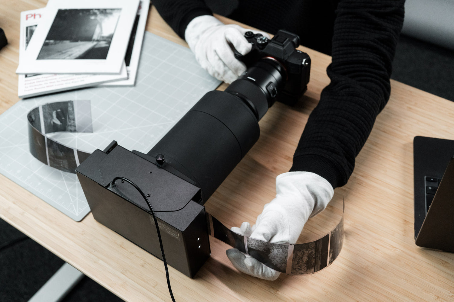 Person handling a digital camera with a camera scanning tool and a roll of film on a wooden table.