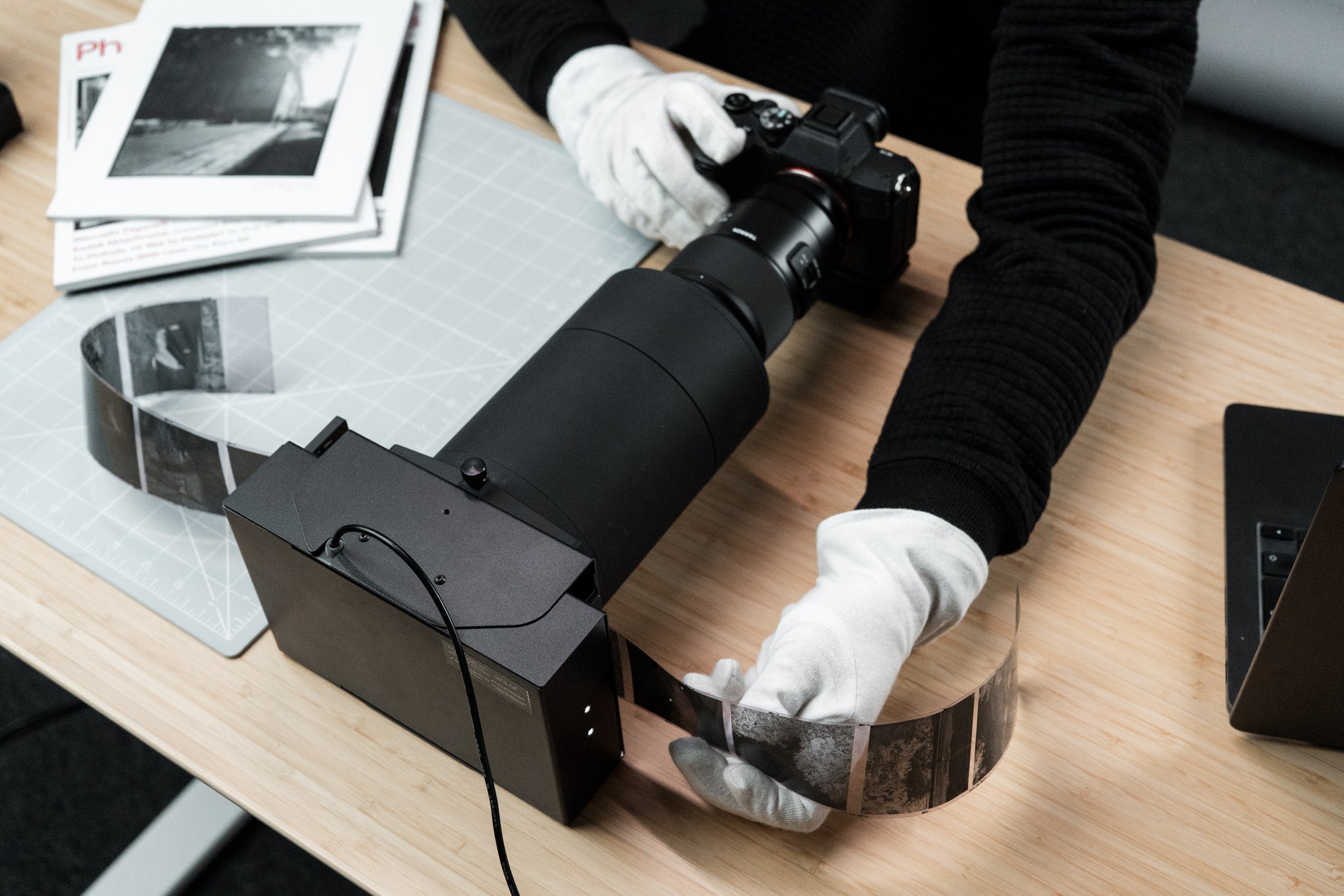 Person handling a digital camera with a camera scanning tool and a roll of film on a wooden table.