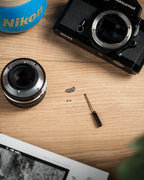 A metallic Nikon camera accessory and screws on a wooden table surrounded by lens and a camera body..