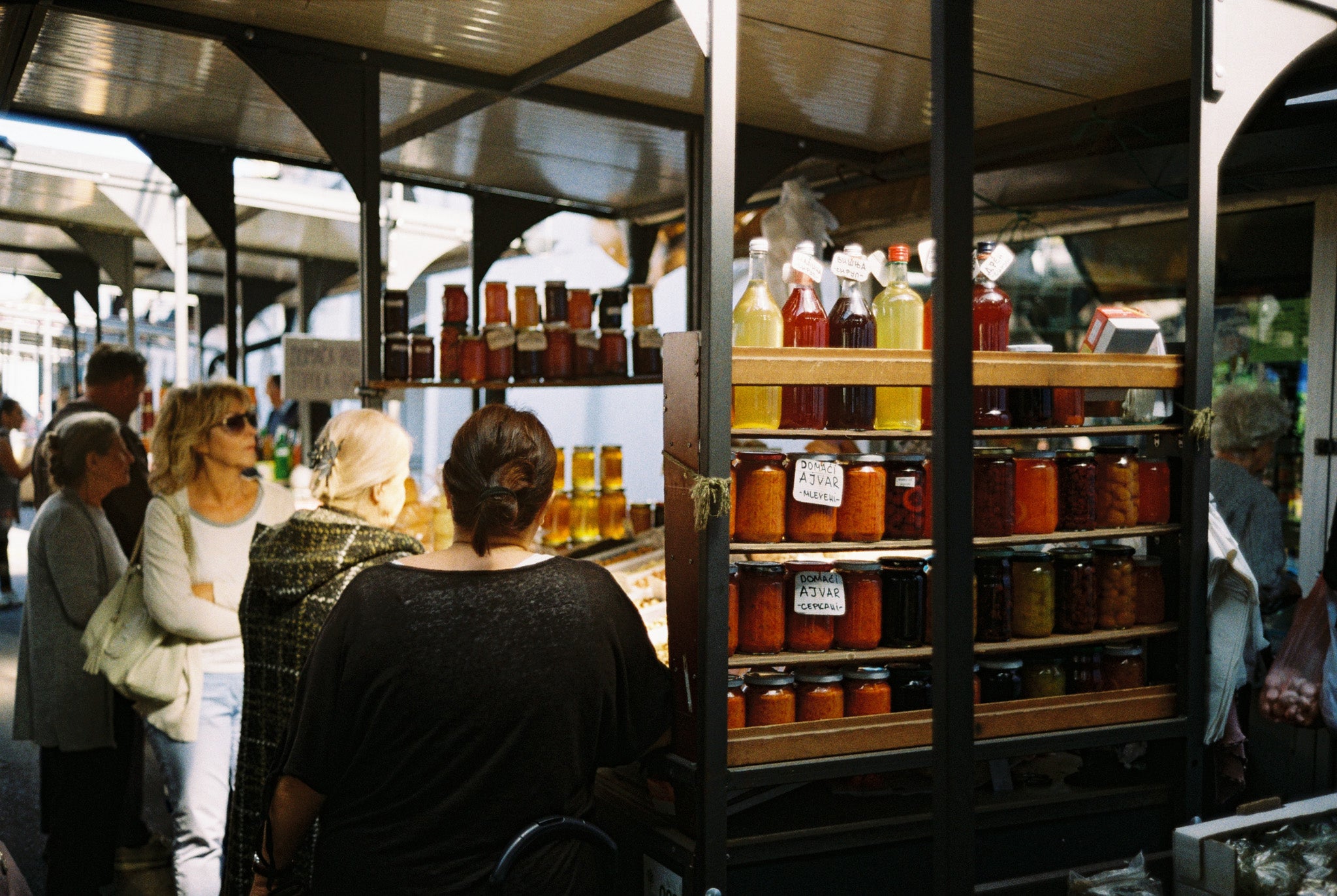 People browsing jars and juices at the market with some market buildings on the background.
