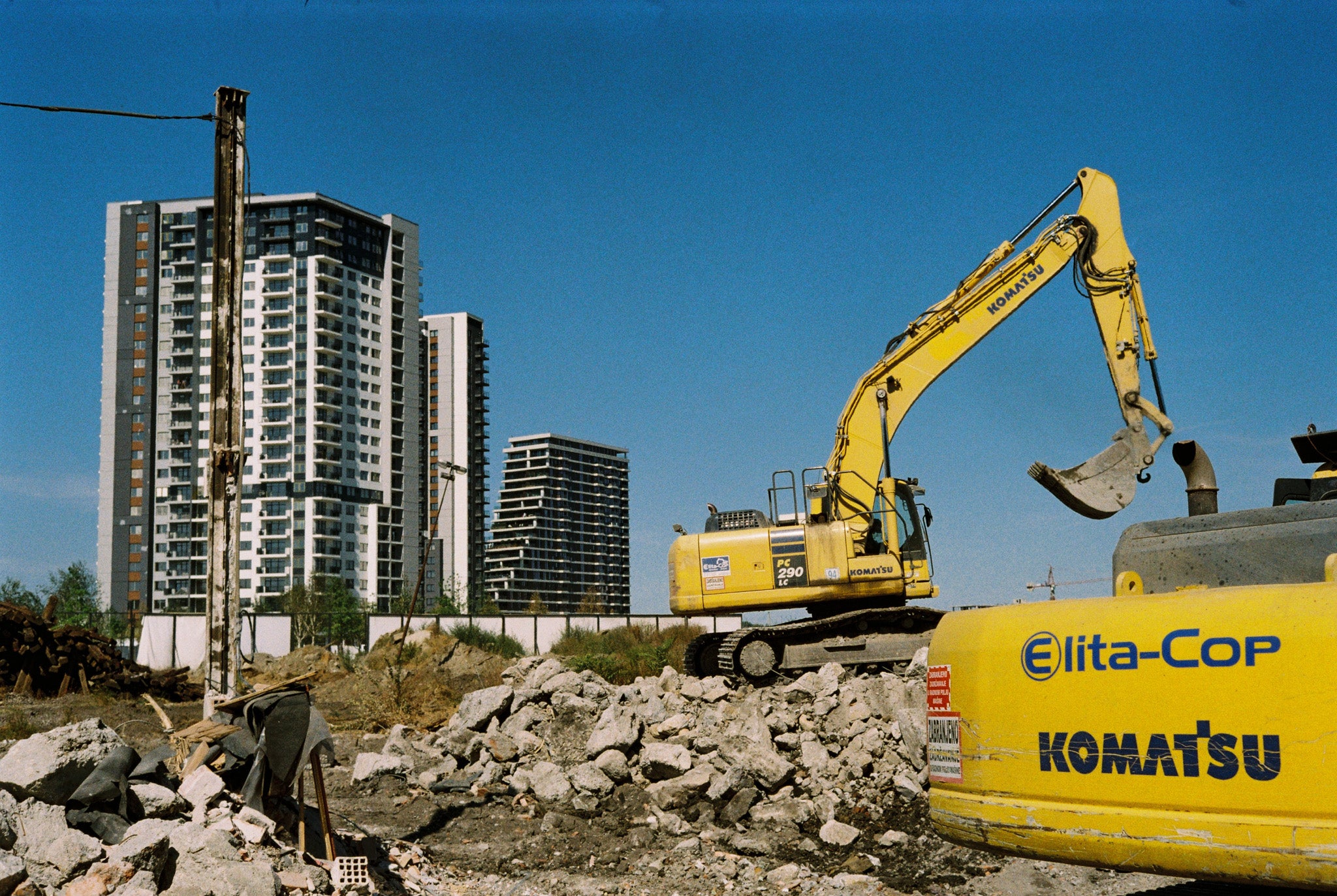 Construction area and excavators in the foreground with some buildings and clear blue sky on the background.