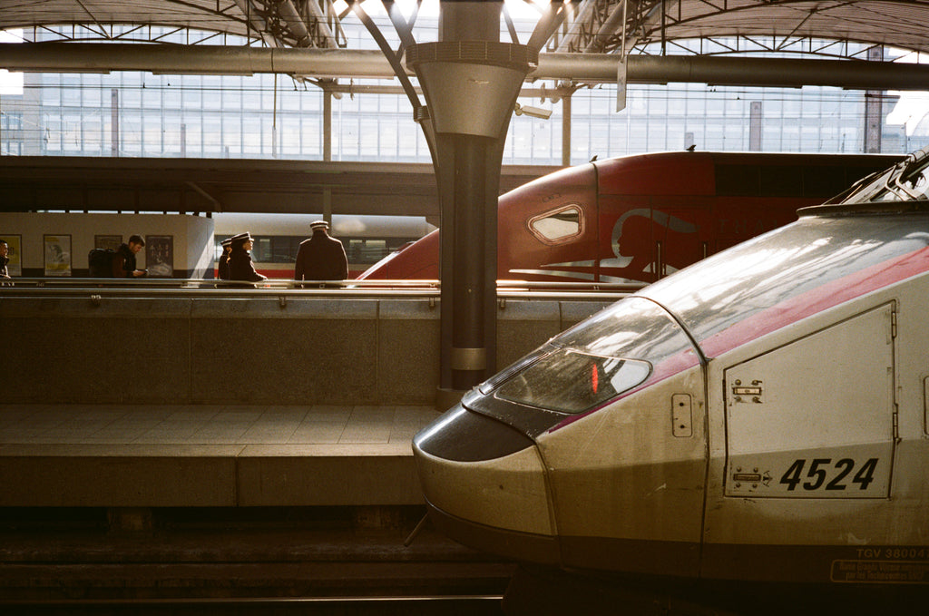 A train station with a train in the foreground and some people, train and windows on the background.