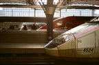 A train station with a train in the foreground and some people, train and windows on the background.
